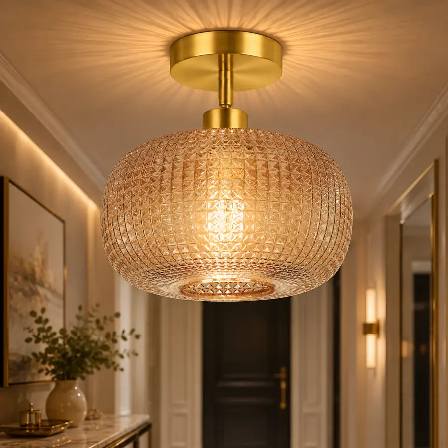 A modern semi-flush mount ceiling light fixture with a textured, faceted amber-tinted glass shade and a brushed gold brass ceiling canopy, shown illuminated in a luxurious hallway setting.