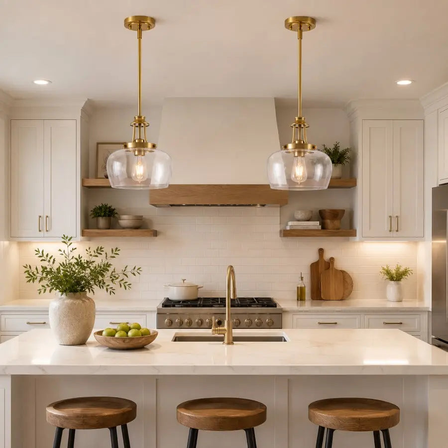 A bright, modern kitchen with two brass pendant lights hanging over a white marble island. The kitchen features white cabinets, a white subway tile backsplash, a gas range, and three wooden bar stools at the island.