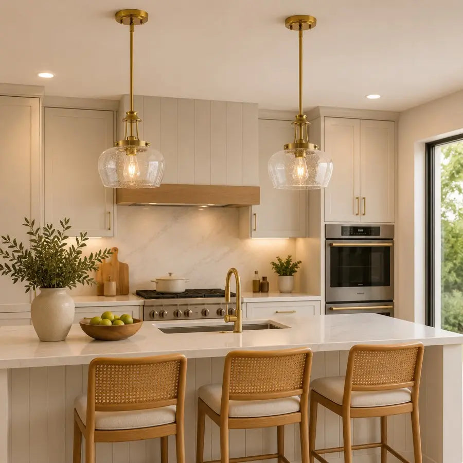 A modern kitchen interior with two brass pendant lights hanging over a white countertop. The space features light wood cabinetry, a built-in stainless steel oven, a marble backsplash, and three woven-back bar stools at the island.