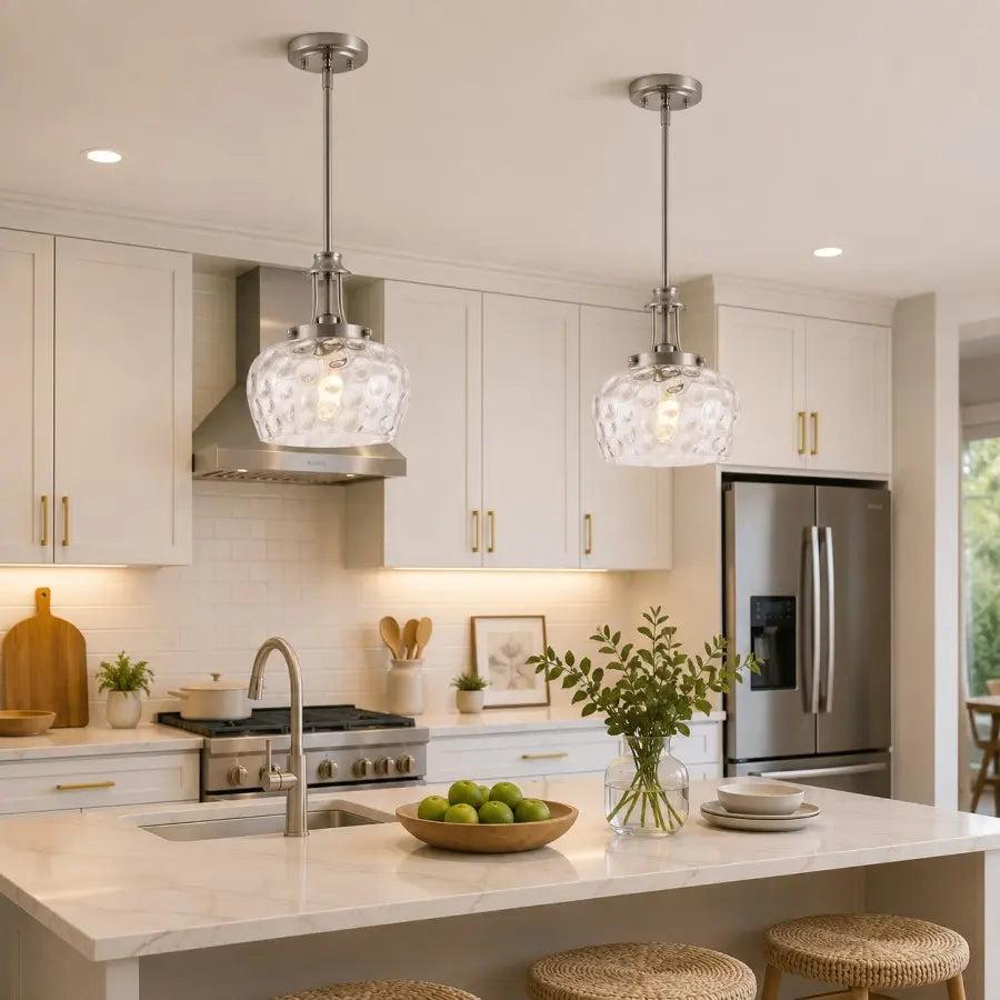 Two modern pendant lights with textured clear glass shades and brushed silver metal rods hang over a white marble kitchen island in a bright, contemporary kitchen with white cabinets and stainless steel appliances.