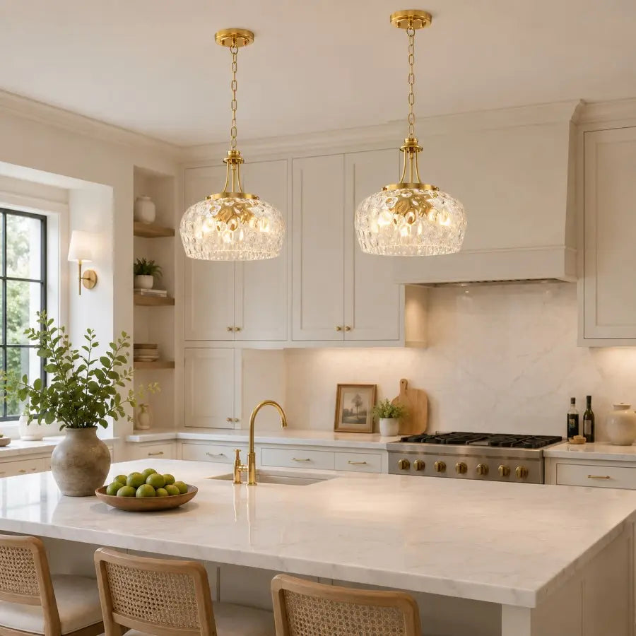 Two gold pendant lights with clear faceted crystal globes hang over a white marble kitchen island in a bright, modern kitchen with white cabinets and a marble backsplash.