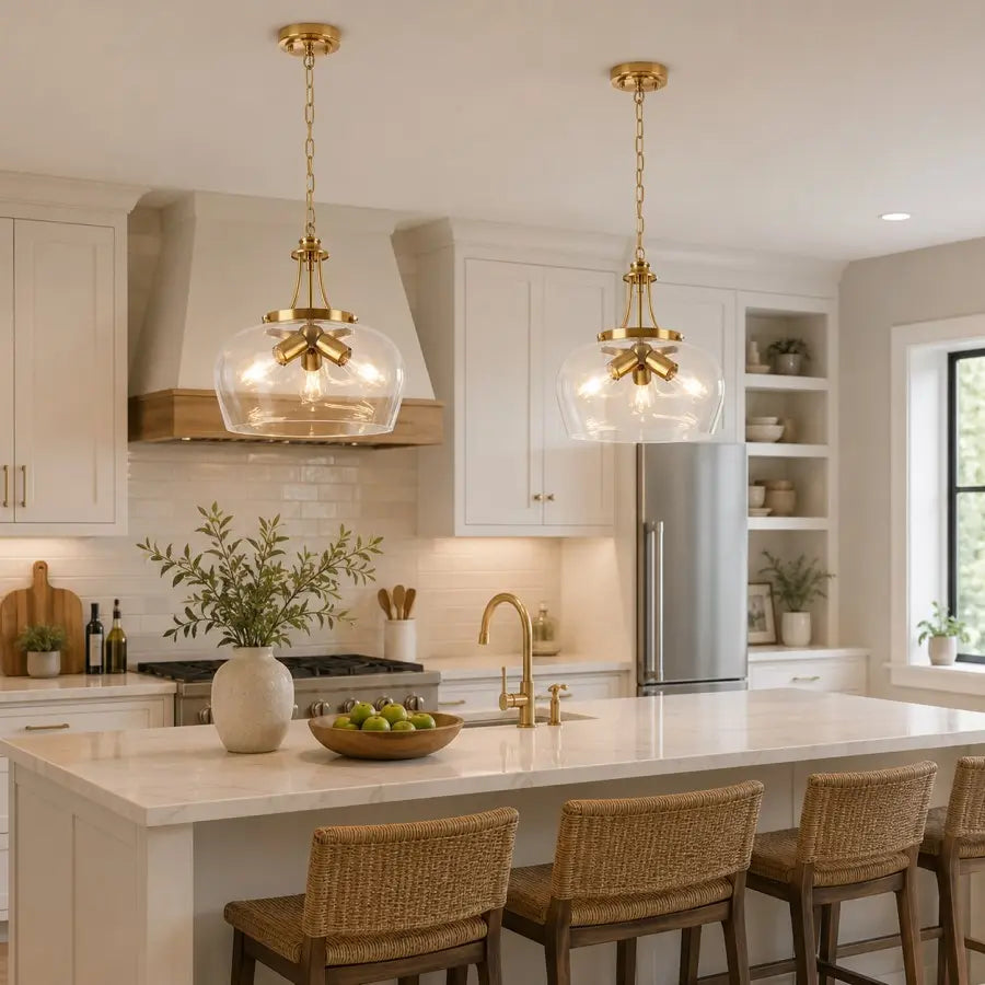 Two brass pendant lights with clear glass globes hang over a white kitchen island in a bright, modern kitchen with white cabinets, a stainless steel refrigerator, and woven bar stools.