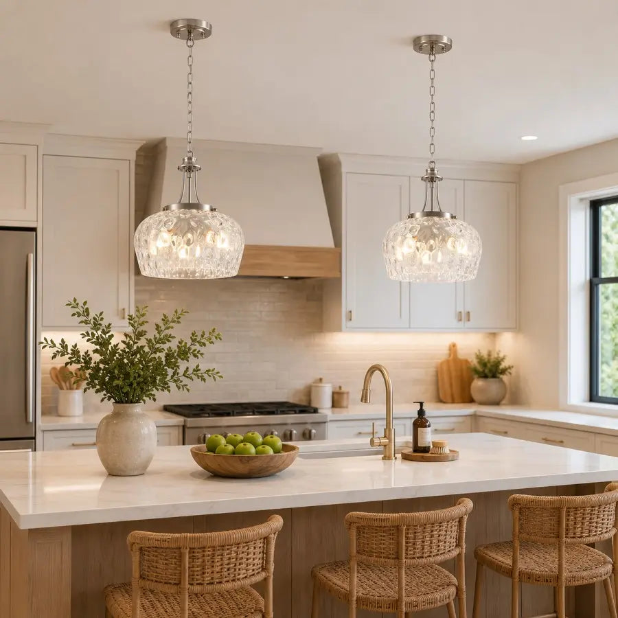 Two modern crystal pendant lights with silver-toned chains hang over a white kitchen island in a bright, contemporary kitchen with beige cabinets, a wood range hood, and woven bar stools.
