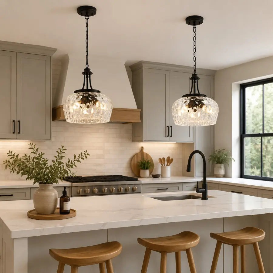 Two black pendant lights with clear textured glass shades hang over a white kitchen island in a modern kitchen with gray cabinets and wooden stools.