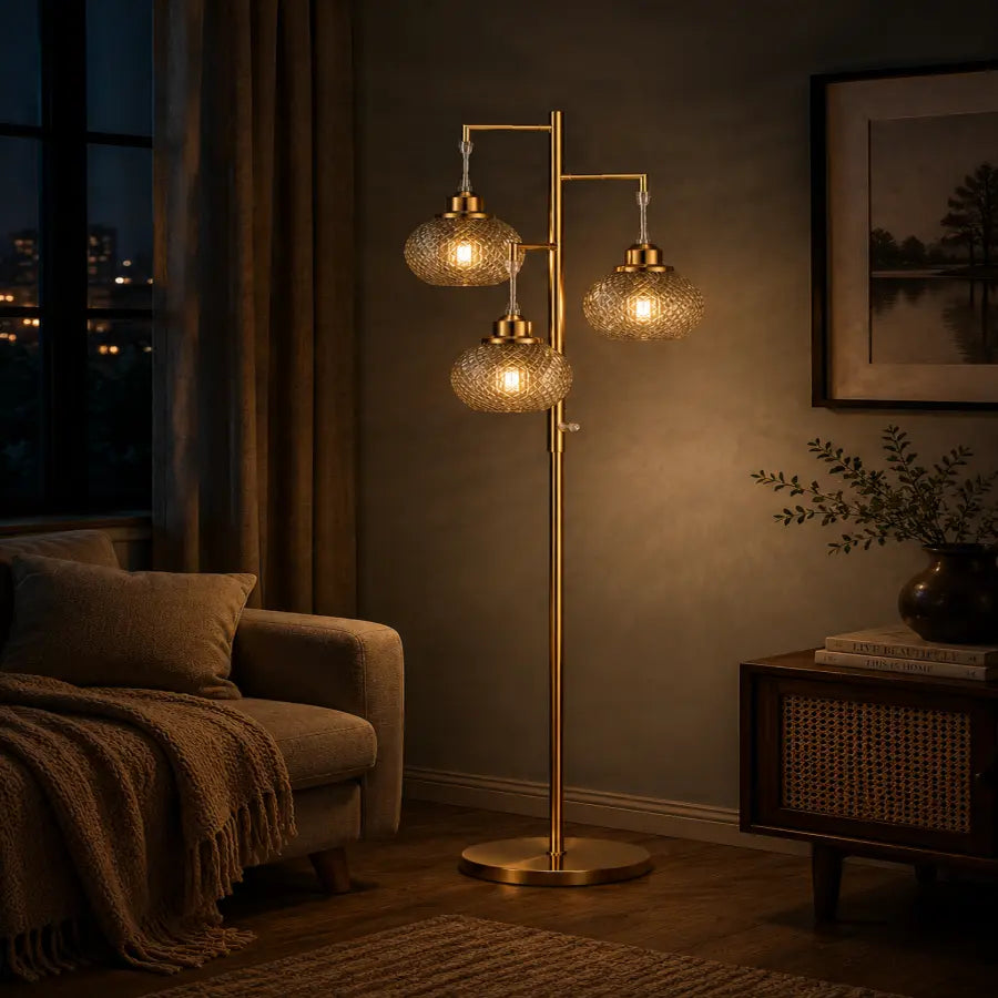 A modern three-light floor lamp with textured glass globes and a brushed brass finish, styled in a cozy, dimly lit living room next to a sofa and a side table.