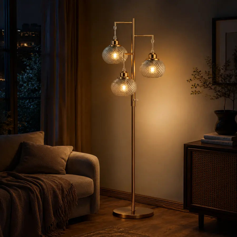 A three-light floor lamp with textured glass globes and a brushed brass stand, illuminated in a cozy living room next to a sofa and sideboard.