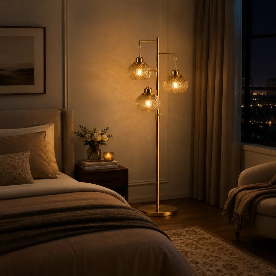 A modern three-light floor lamp with textured glass globes and a brushed brass finish stands in a warmly lit bedroom next to a bed, casting a soft glow in the night.