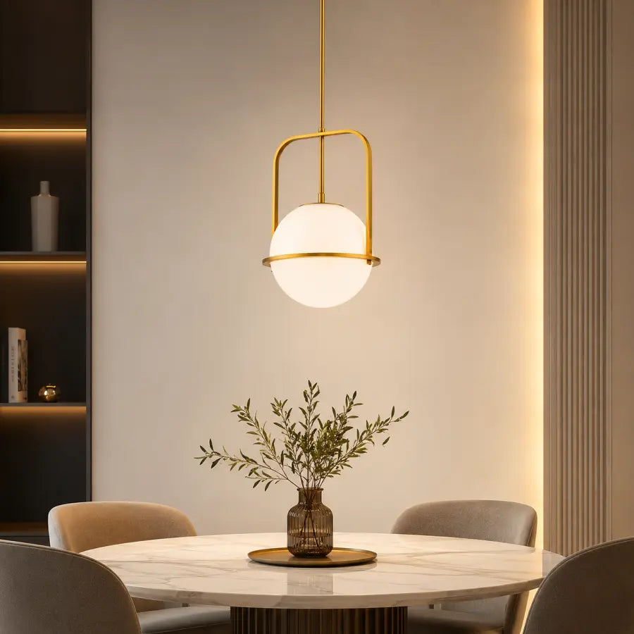 A modern pendant light with a gold metal frame and a frosted white globe shade hangs over a round marble dining table with chairs and a vase of greenery in a warm, contemporary interior.