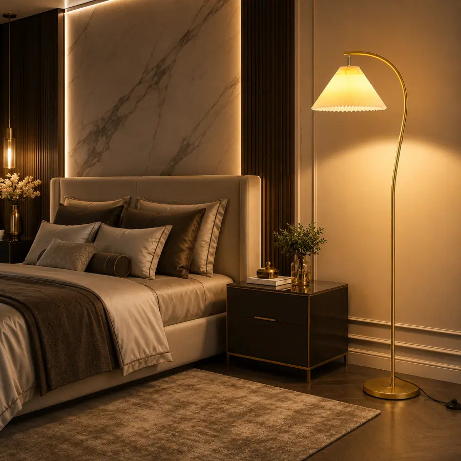 A modern bedroom scene with a bed, bedside table, and a tall floor lamp with a warm glowing shade and curved gold-toned stand. The room has warm beige walls, dark wood paneling, and a marble accent wall behind the bed.