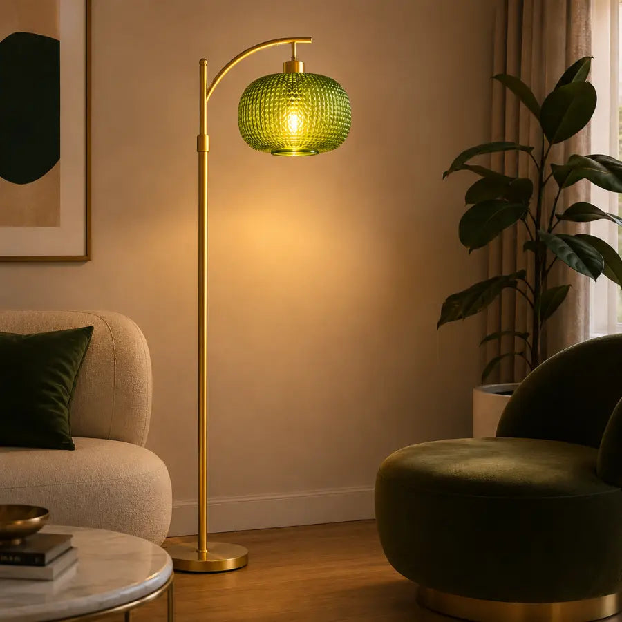 A modern floor lamp with a gold-finished metal stand and a textured green glass shade glowing with warm light, placed in a styled living room next to a sofa and plant.